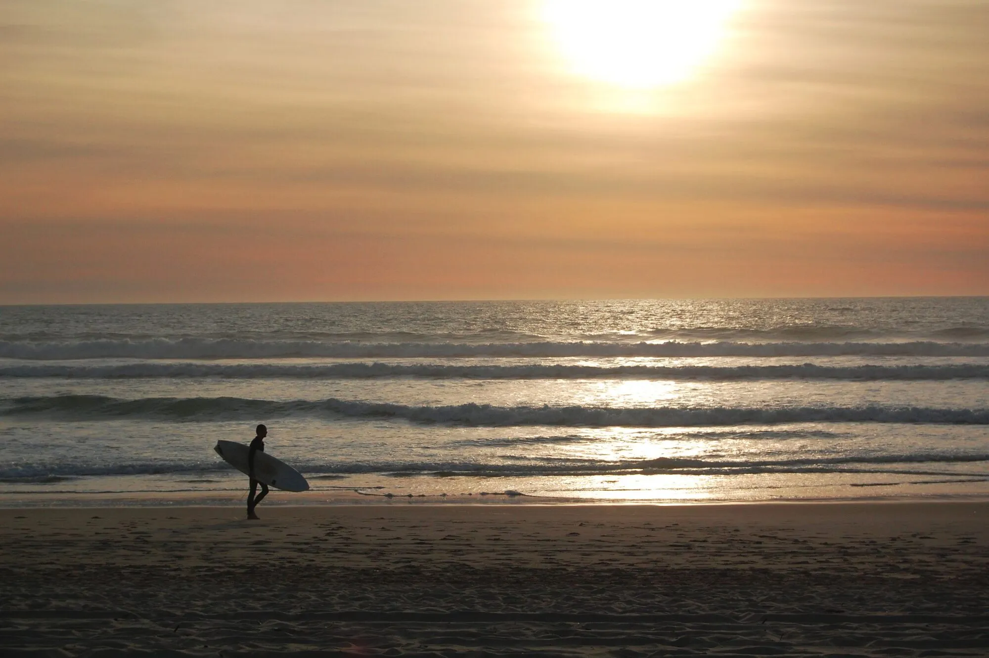 A surfer walking back across the sand at Mimizan, last light on the water